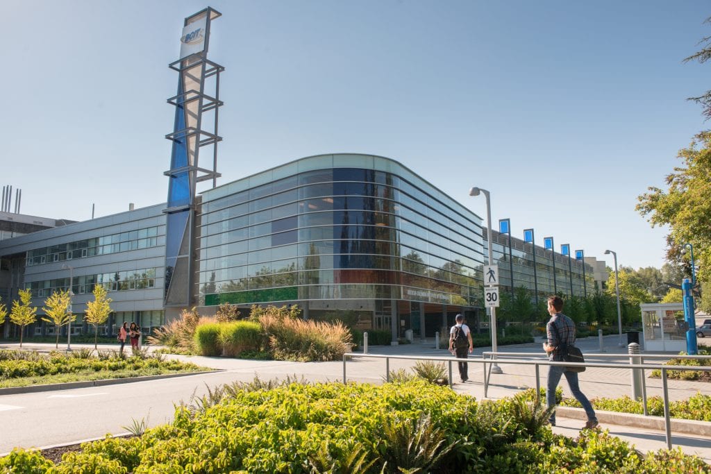 Burnaby campus SW01 building with 2 students walking towards it and 2 students standing outside of it..