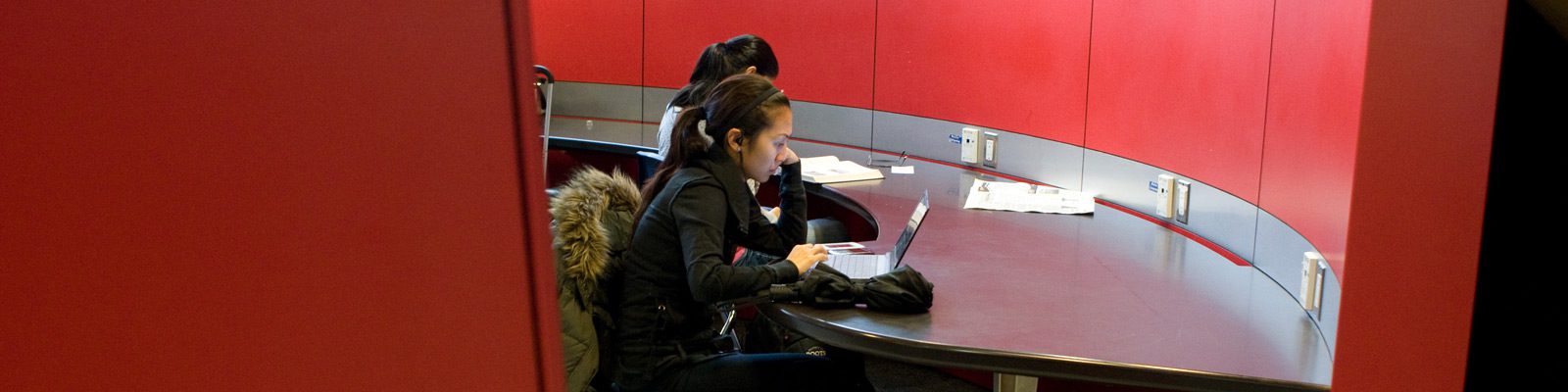 two students looking at laptops in a red walled room on a silver table.