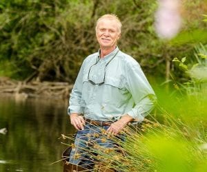 man with grey hair and mustache stands in a creek wearing waders and has glassing hanging around his neck, hands on hips and smiling