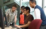 Teacher explaining something to 3 students located in front of a computer terminal.