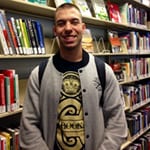 Man smiling with short brown hair, wearing a grey button up jacket in front of library books.