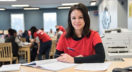 Female tutor seated at a desk wearing red shirt and has shoulder length brown hair.