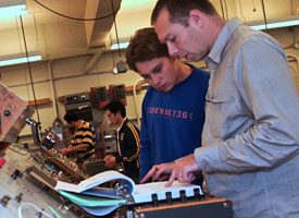 Two men reading specs in front of a mechanical panel of knobs.