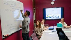 Instructor writing on a white board with 3 students seated at desks observing.