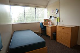 Image of an on-campus housing room with a bed, dresser, desk, chair and desk lamp.