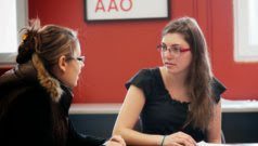 Two ladies with long brown hair talking to each other. Both have glasses.