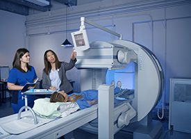 Photo of two students operating a nuclear medicine machine for a patient lying on a bed.
