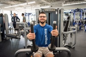 man sitting on excercise equipment wearing a BCIT recreation T-shirt and smiling
