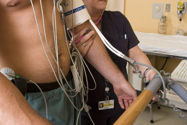 A healthcare worker monitors a man on a treadmill stress test, attaching electrodes to his chest and using a blood pressure cuff.