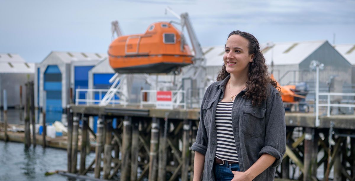 Woman standing at the harbour, with a lifeboat docked up high in the background.