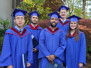 Five graduates from the Technology Management BTech program, attending BCIT June 2023 graduation ceremony, stand outside together dressed in blue BCIT Convocation gowns against a backdrop of a grey campus building, and green and brown foliage.