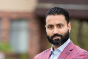 TMGT grad, Sunny Jassal, standing outside brick building, wearing a pink and blue jacket, as featured in Drishti Magazine