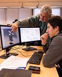 Male instructor with grey hair pointing at a computer where a male student is sat.