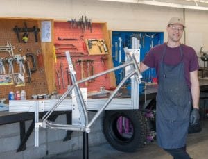 young guy with baseball cap and apron stood beside a bicycle frame