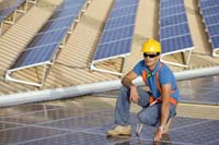 man kneeling down in sunglasses, blue jeans and yellow hardhat - solar panels in the background