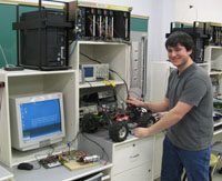 male student with brown hair in a grey t-shirt at a desk with a car-like robot and computer