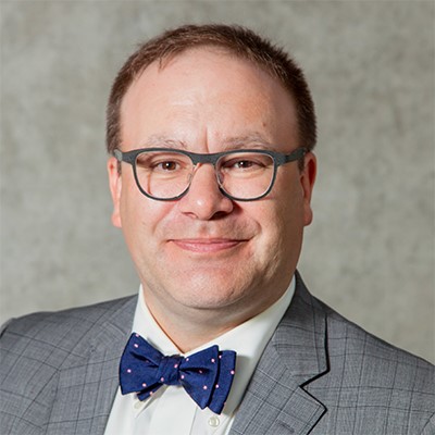 Brett Ballah with blue framed glasses, grey suit, and blue bowtie