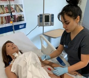 A female nursing student in blue scrubs attends to the donated "Manikin."