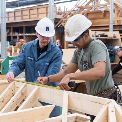 Two men in hard hats in the carpentry trades shop, measuring a length of timber.