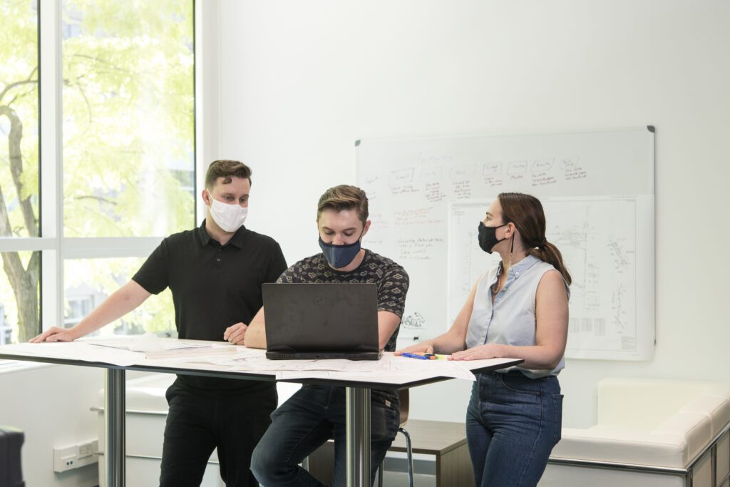 three students with masks on inside a classroom conversing