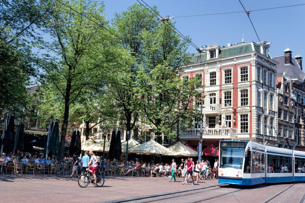 Amsterdam Leidseplein with cyclists, pedistrians, a tram and a full outdoor patio in front of a 5 story brick building.