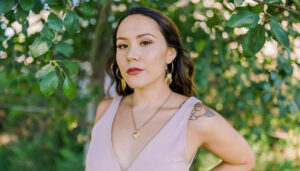 Diosa Femme with shoulder length brown hair, gold earrings, gold necklace, red lipstick, tattoo on the shoulder, and a pink blouse