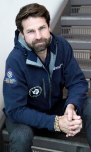 Eric with beard, blue winter jacket and sitting by the staircase