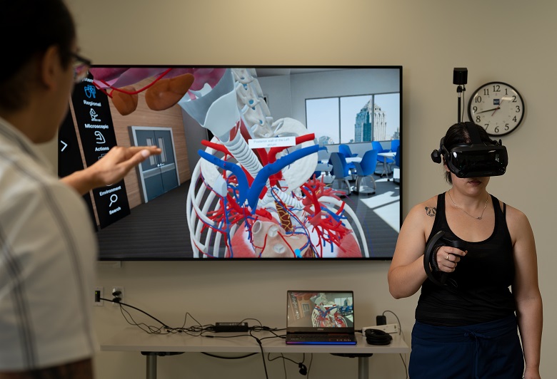 Woman wearing VR headset, person directing action, screen in background showing anatomy