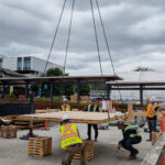 BCIT MaTT Hub participants in high-visibility vests guide a large CLT floor panel being lowered by crane chains onto a concrete slab.