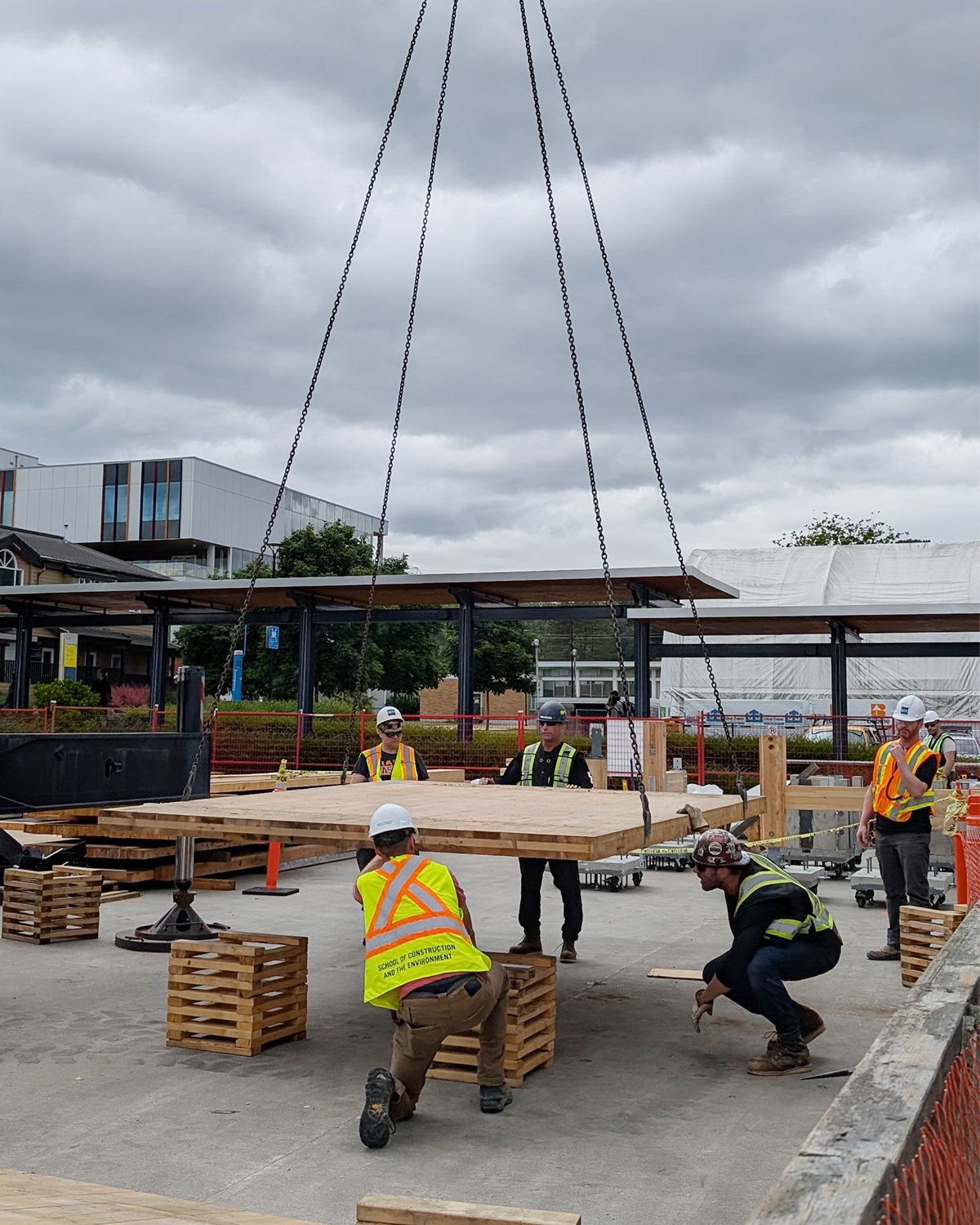 BCIT MaTT Hub participants in high-visibility vests guide a large CLT floor panel being lowered by crane chains onto a concrete slab.