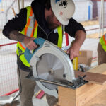 A student wearing a BCIT high-visibility vest uses a circular saw to cut a mass timber component on the practicum site.