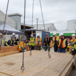 An instructor addresses the full student group gathered around a large CLT panel rigged with chains, briefing them before a crane lift exercise on the BCIT campus.