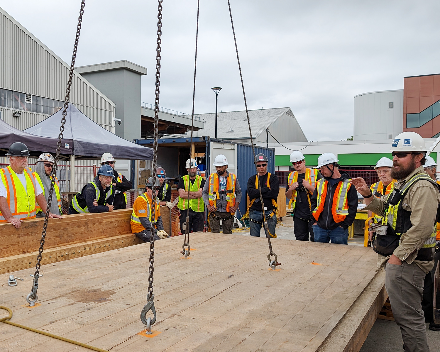 An instructor addresses the full student group gathered around a large CLT panel rigged with chains, briefing them before a crane lift exercise on the BCIT campus.