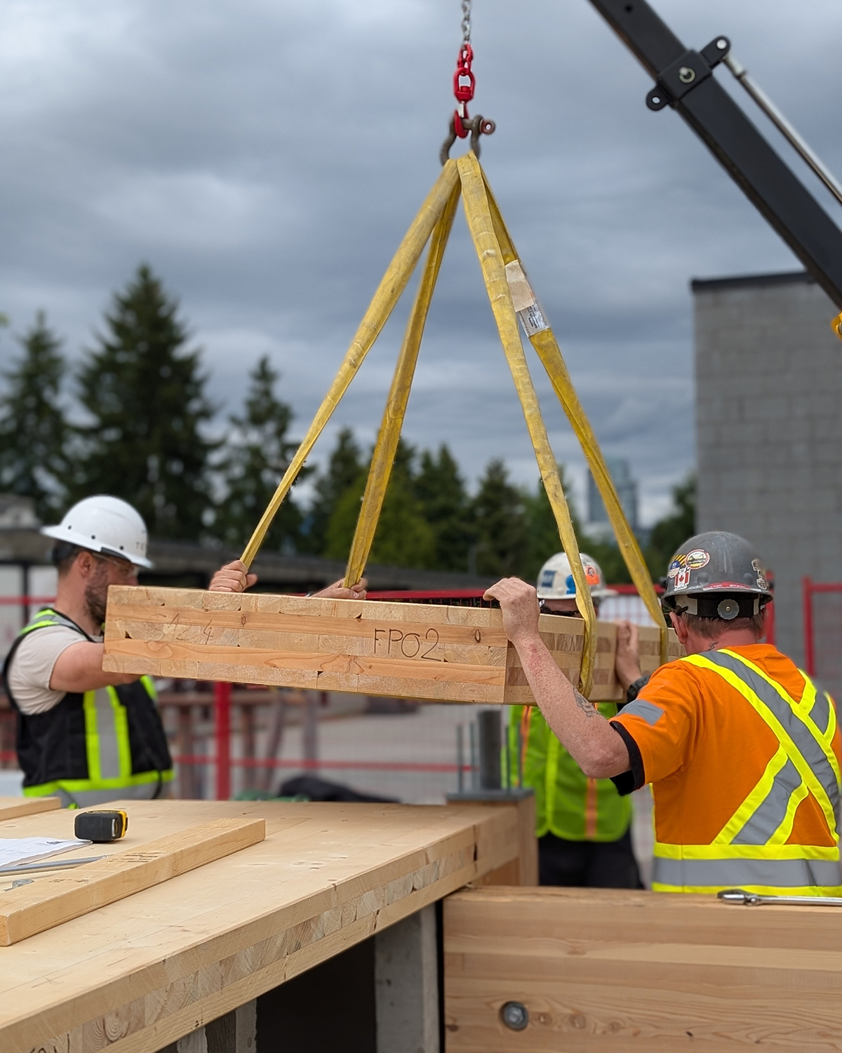 A group of MaTT Hub students in safety gear work together to position a labelled glulam beam suspended by rigging straps from a crane arm.