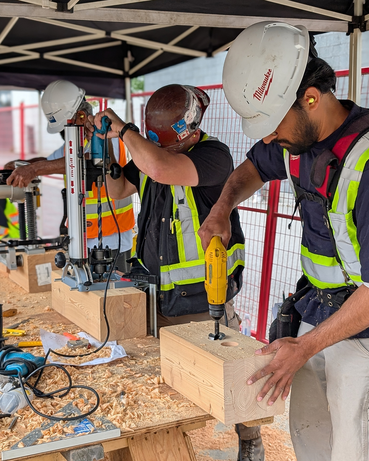 Three students in hard hats drill into mass timber connection blocks using power tools during practicum training.