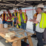 A BCIT instructor leads a small group of practicum participants through a hardware demonstration, showing mass timber connection hardware and fasteners laid out on a workbench.