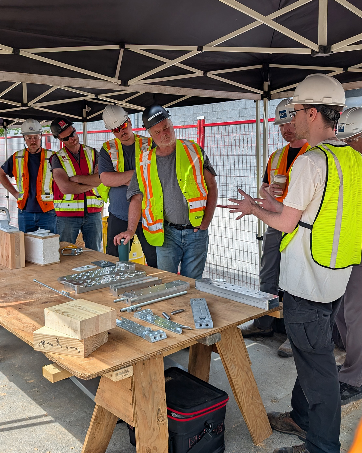 A BCIT instructor leads a small group of practicum participants through a hardware demonstration, showing mass timber connection hardware and fasteners laid out on a workbench.