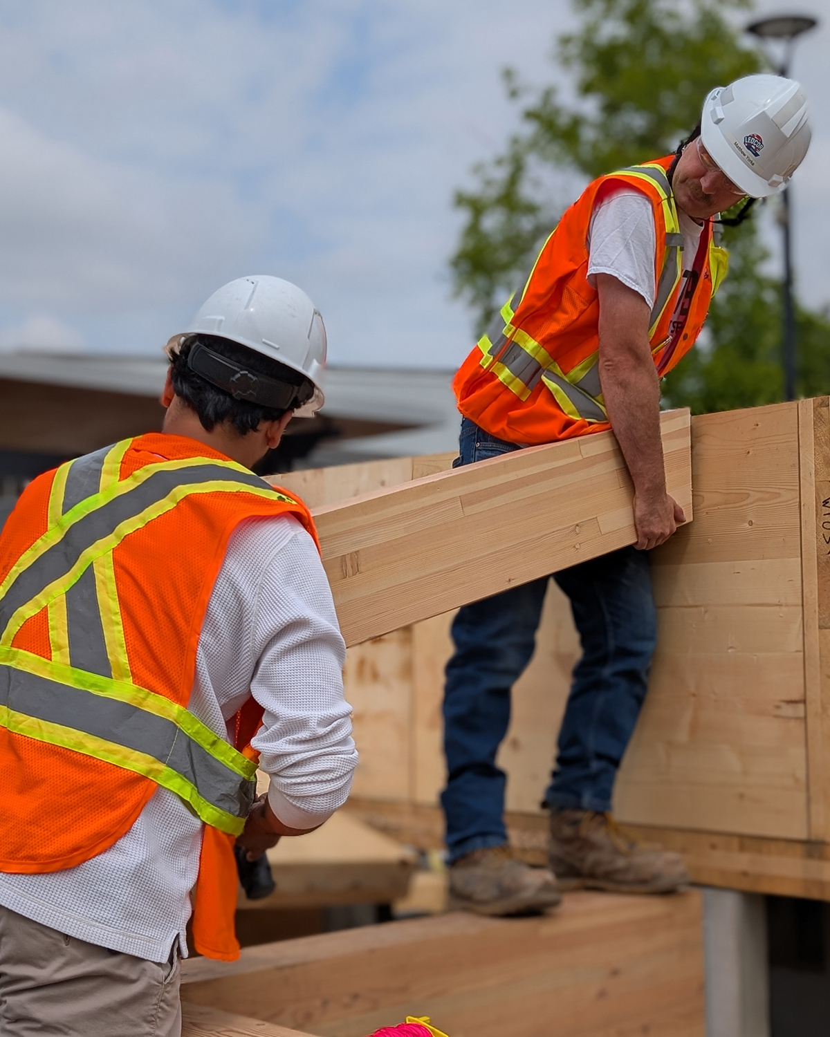 Two students in high-visibility vests and hard hats manoeuvre a glulam floor plank into position on top of a mass timber wall assembly.