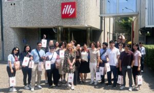 Group of students smiling and standing together in front of an Illy Café company sign, capturing a moment during their study abroad experience