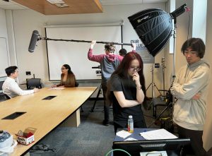 Students observe a scene on screen while filming takes place in the background.