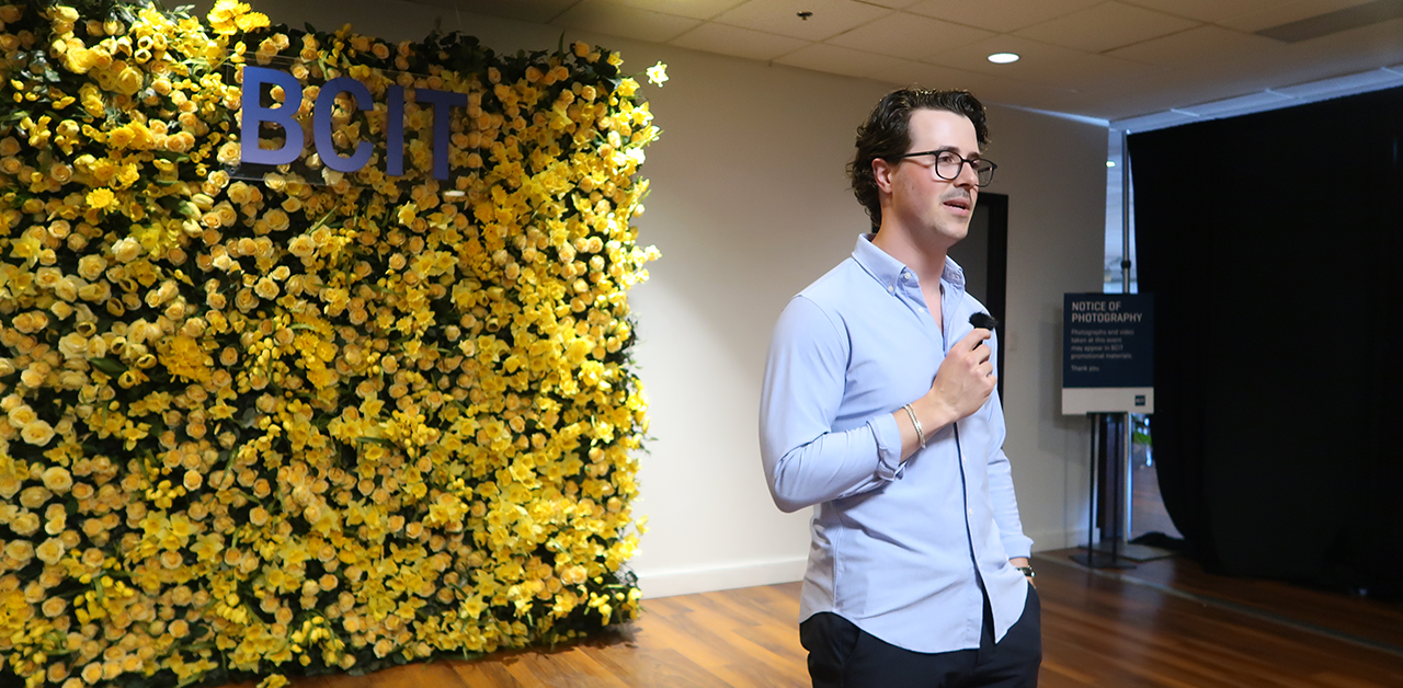 Matt Cox delivers a speech at the BCIT 2024 Spring Convocation Graduating Awards Ceremony. He holds a microphone, and stands in front of a yellow flower wall with a sign on it that reads "BCIT".