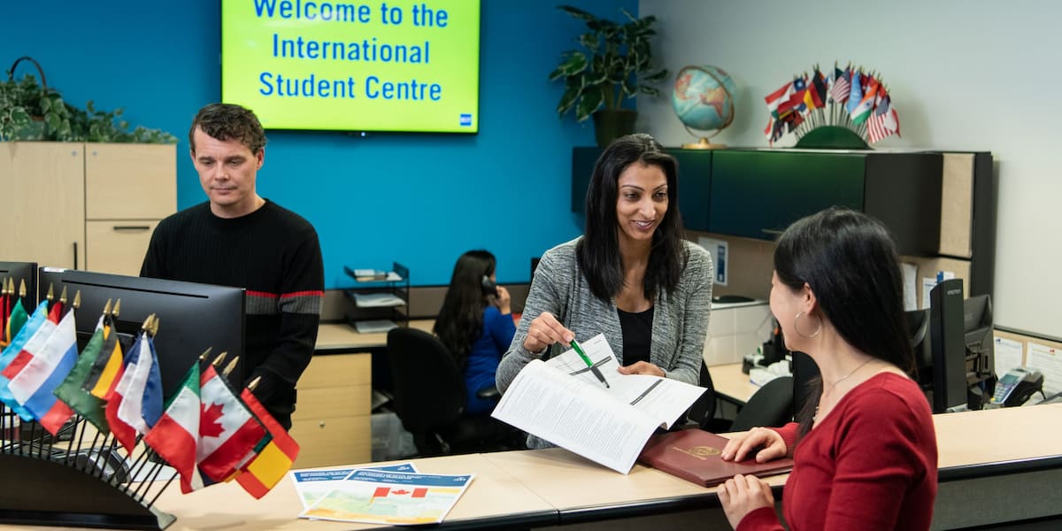 An image of the International Student Centre front desk. There is a woman showing a student a document, a man working on a computer, and a woman speaking on the telephone in the background.