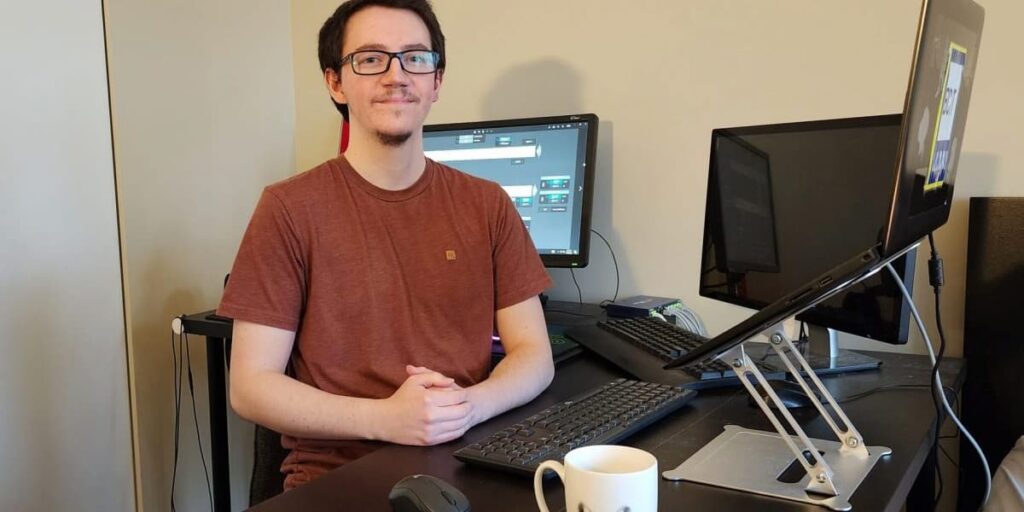 man with brown shirt and glasses sits at workstation with three screens and two keyboards