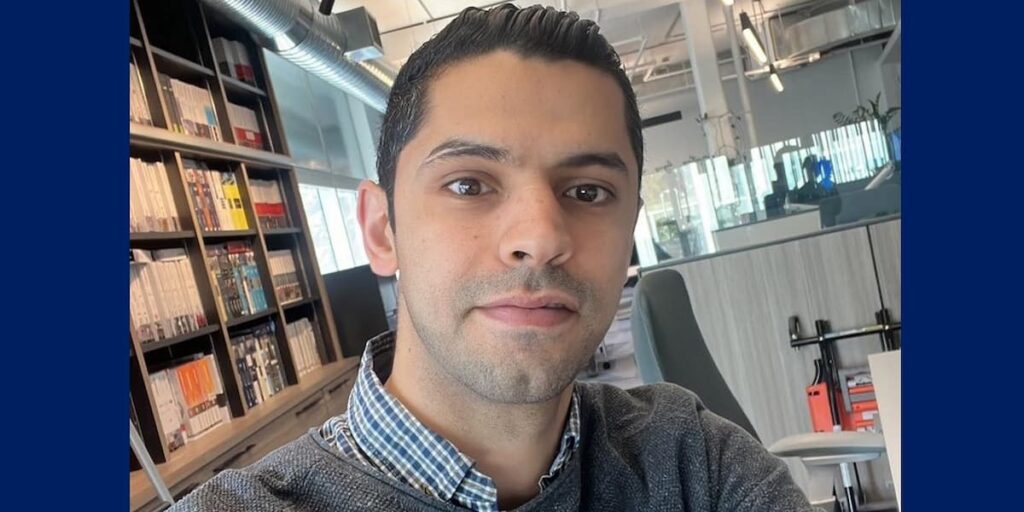 Man with short dark hair wears a grey sweater with a collared check shirt in an office with bookcase in the background