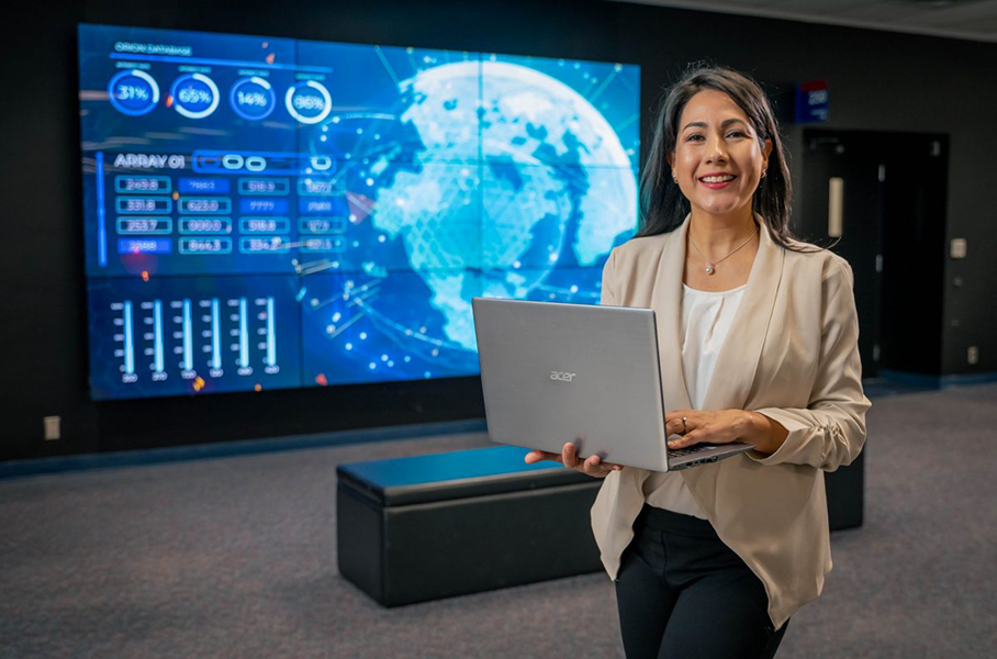 Smiling woman with long brown hair in a light blazer holds a laptop while standing in front of a screen displaying data and graphs