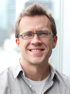 White man with glasses in a grey shirt smiles in front of a downtown background