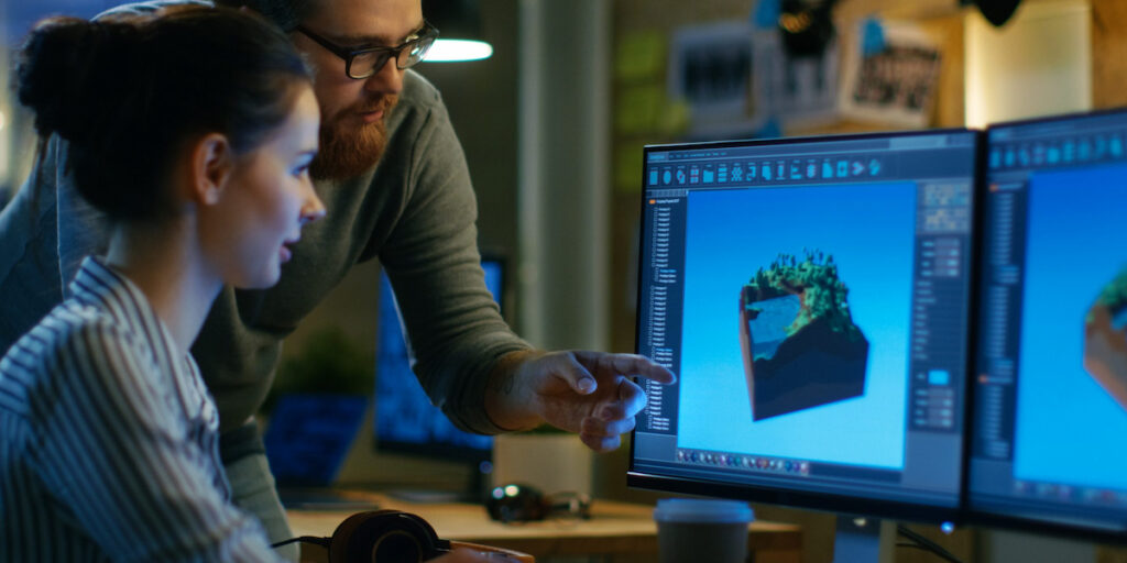 Female Game Developer Has Discussion with Male Project Manager While Working on a Game Level on Her Personal Computer with Two Displays. They Work in a Modern Loft Office Creative Environment