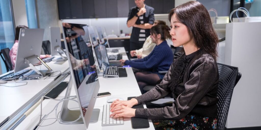 A focused woman working at a desktop computer in a modern computer lab, with several other people seated in a row using similar workstations. A man stands in the background observing or instructing. The setting is bright, organized, and professional, with large monitors and minimalist design.