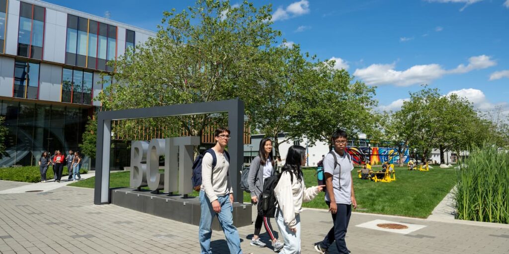 Four students walking past a large outdoor BCIT sign with greenery and modern buildings in the background.