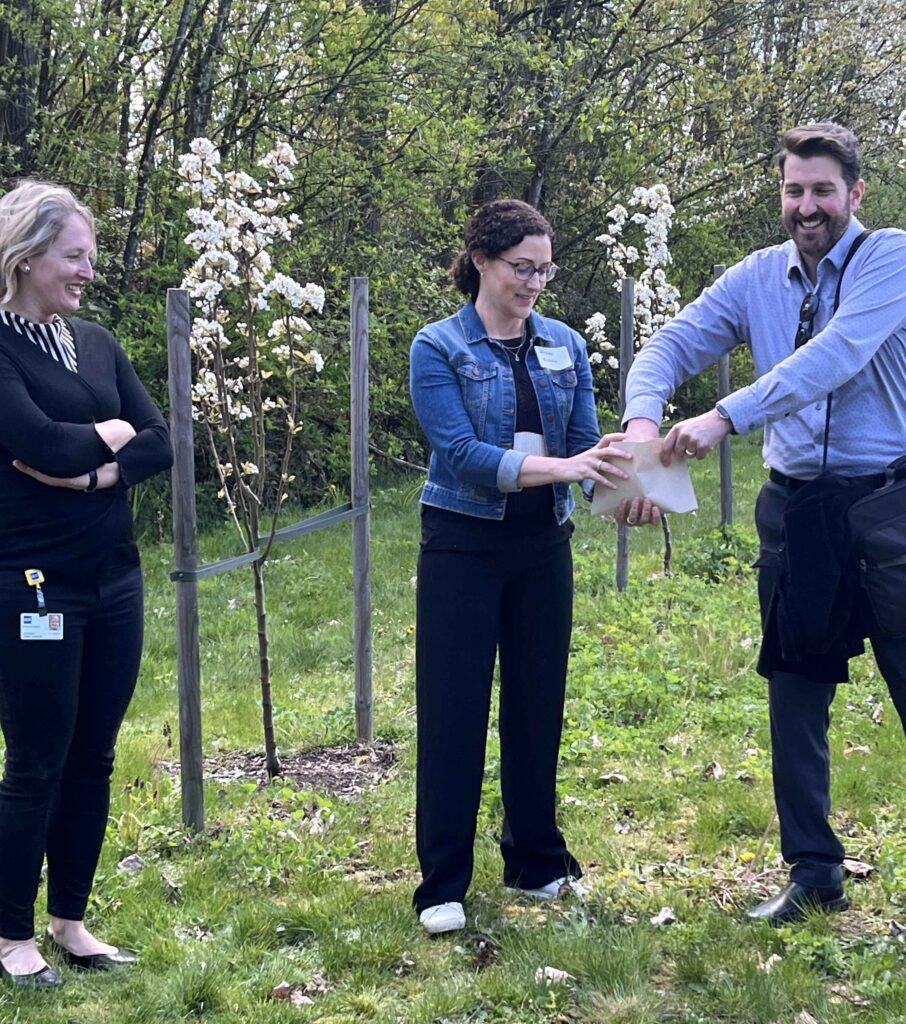 three people stand in front of a small flowering orchard while drawing prize names from an envelope
